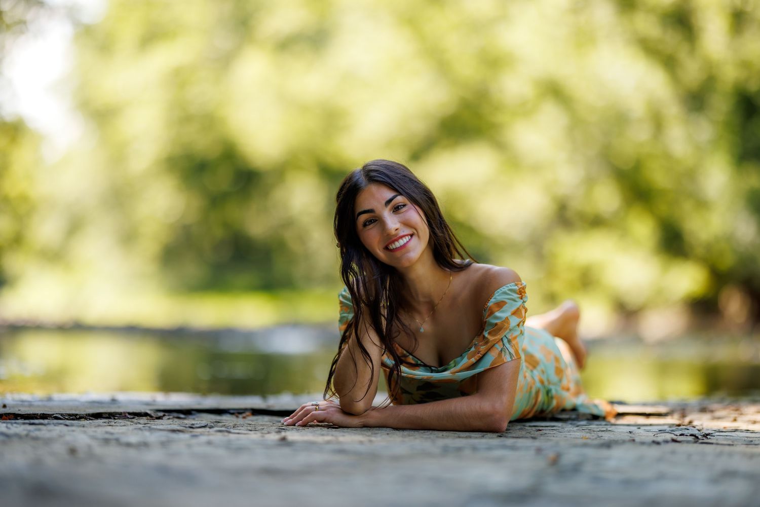 Smiling brunette woman in a floral off-shoulder dress lying on a stone surface near a sunny river.