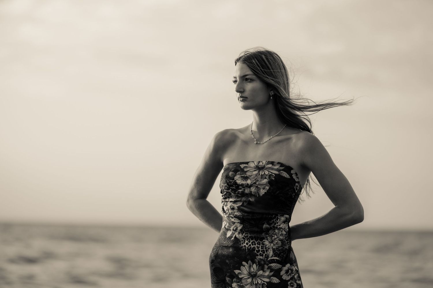 Black and white portrait of a woman in a strapless floral dress standing on a beach with wind in her hair.