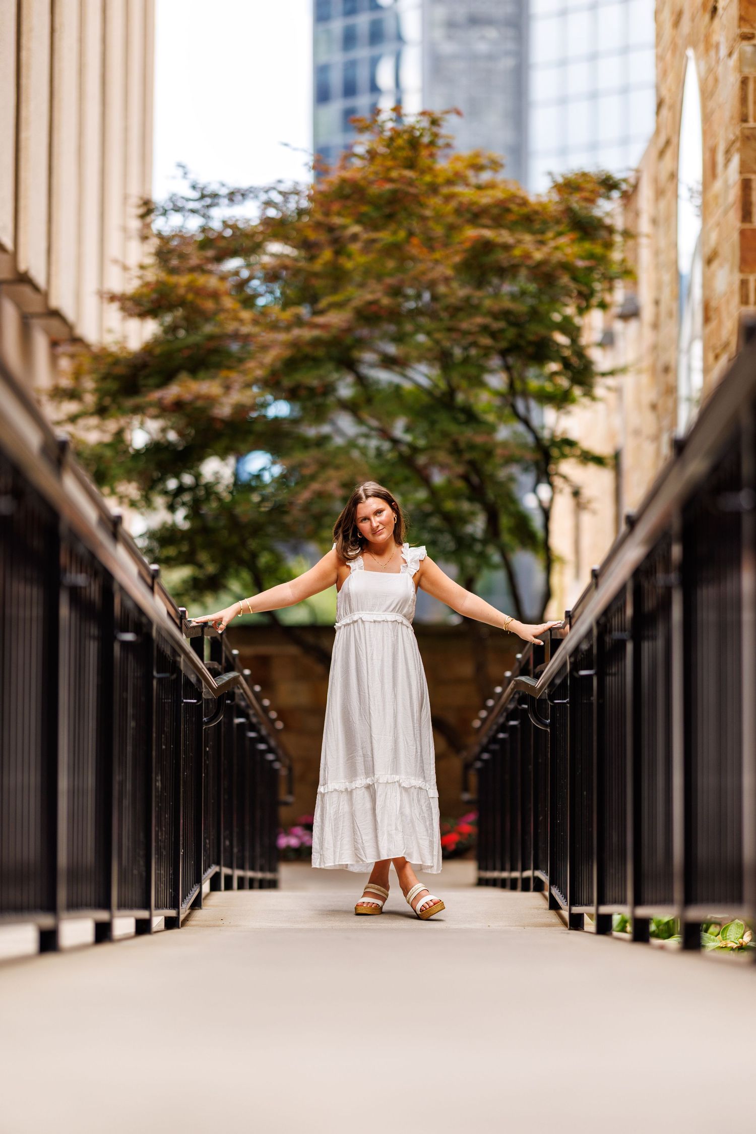 Woman in white sundress standing on urban walkway with arms outstretched between railings.