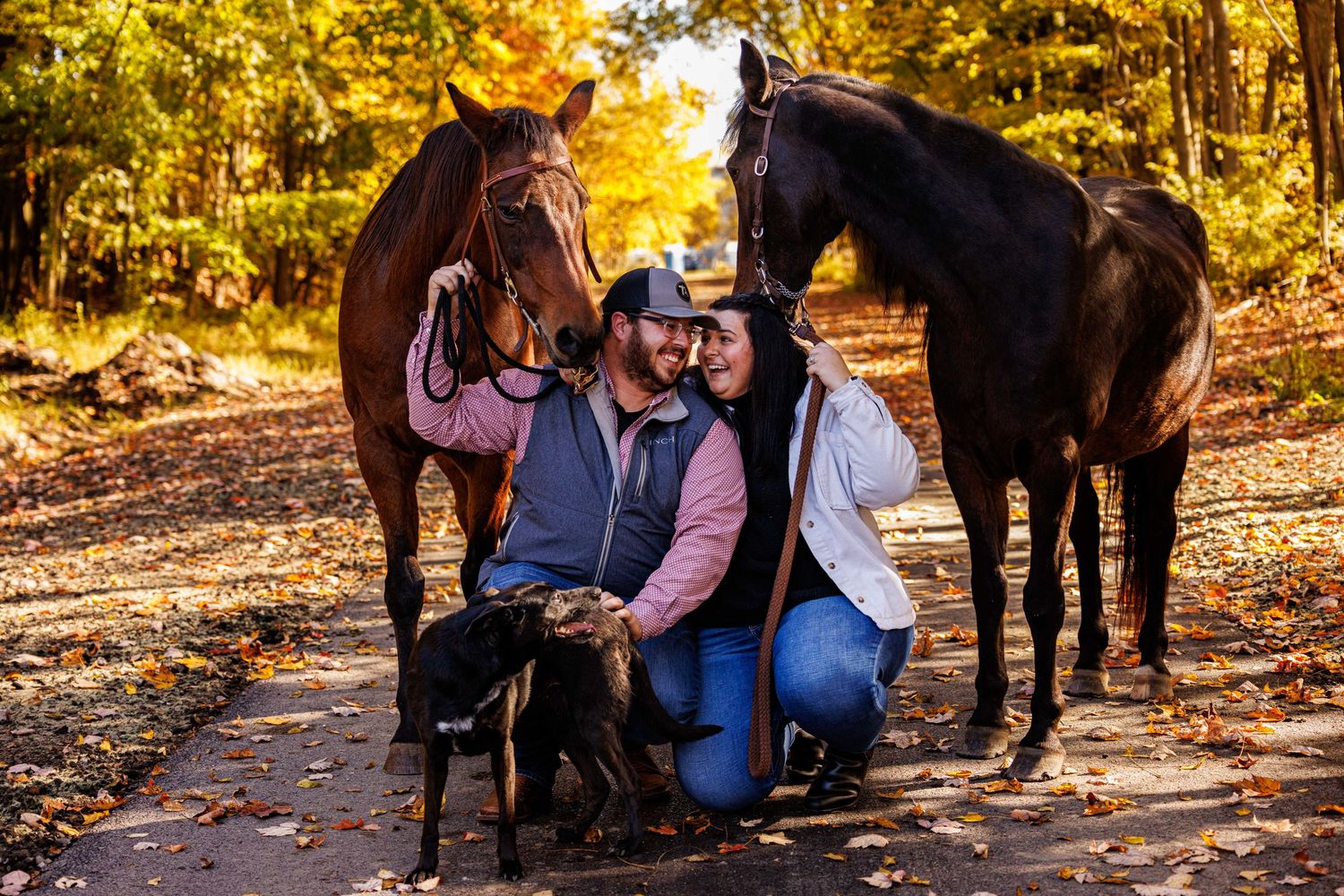 A smiling couple kneels together with two horses and a dog among autumn foliage.