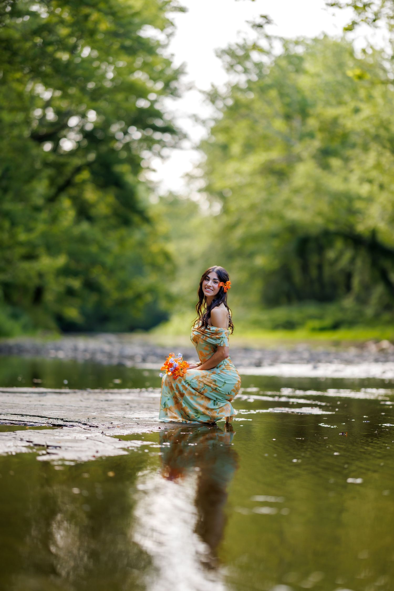 Smiling woman in a colorful dress holding flowers while standing in a shallow tree-lined river.