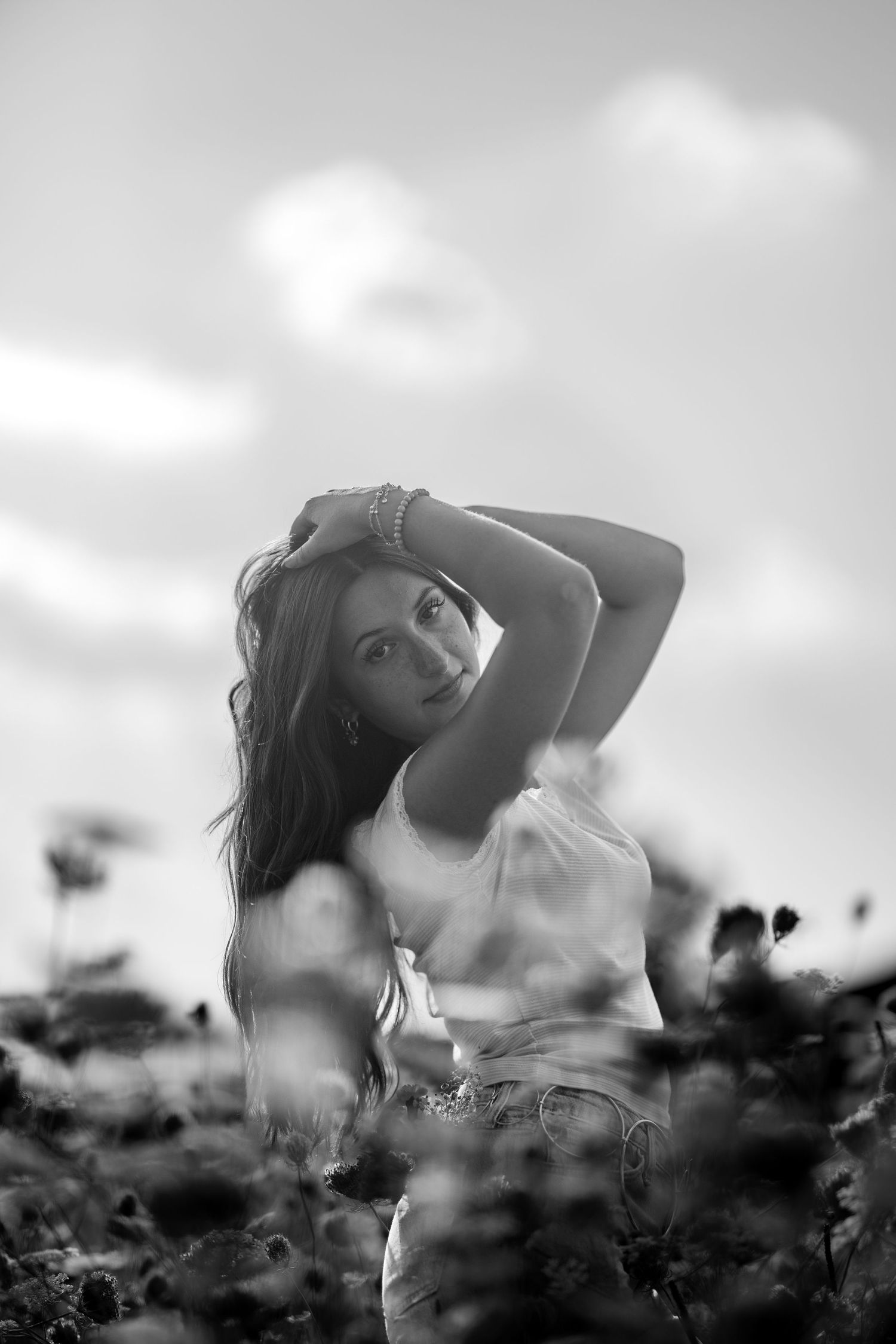 Black and white portrait of a young woman with arms raised posing in a wildflower field under cloudy sky.