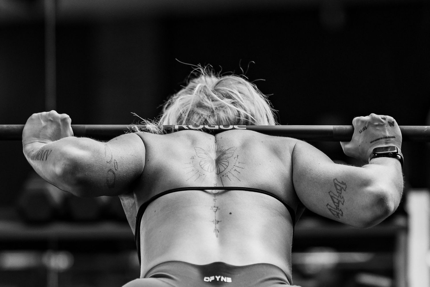 Black and white rear view of muscular female athlete performing pull-ups on bar with tattoos visible.
