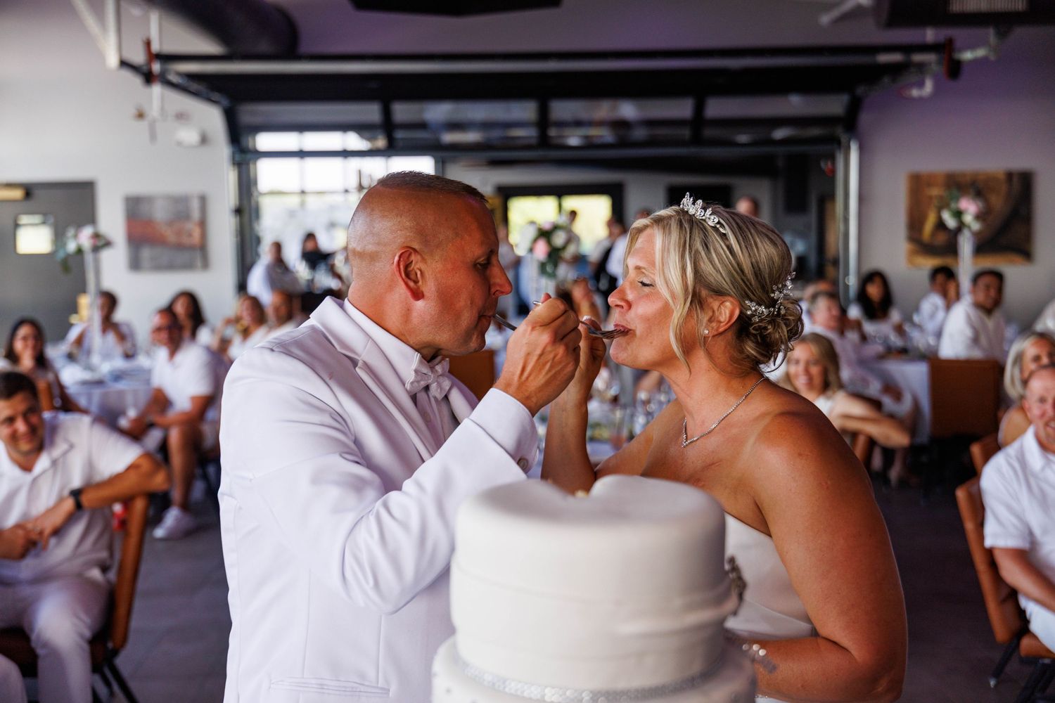 Groom feeds bride a piece of wedding cake during their reception as guests watch.