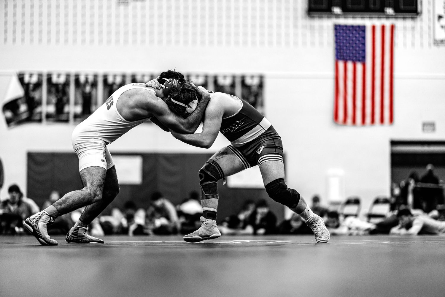 Two wrestlers engage in a grappling match on the mat with an American flag displayed in the background.