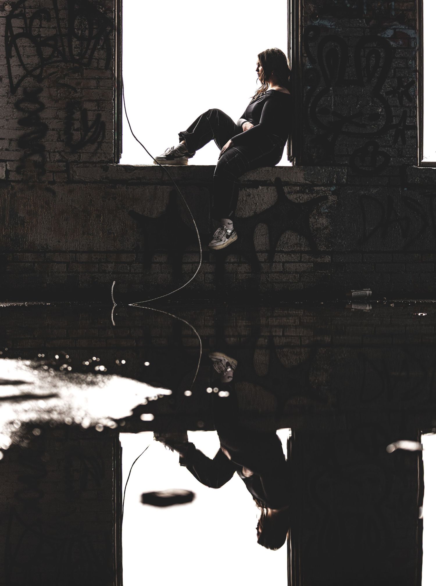 Woman sitting in abandoned building window with fishing rod perfectly reflected in water below.