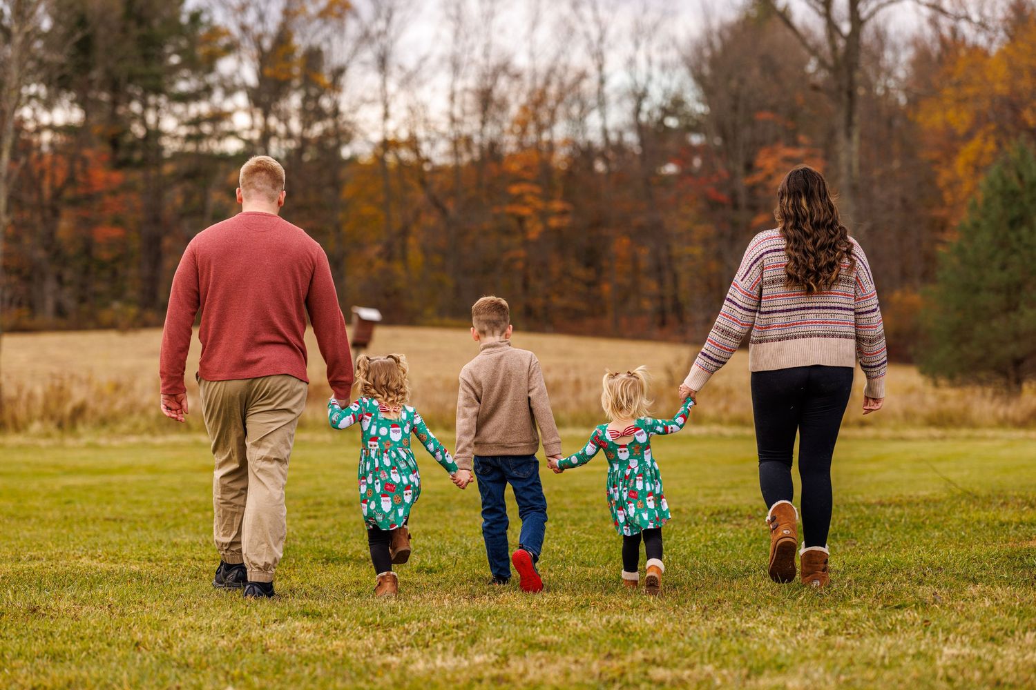 Family of five walking hand in hand across a green field with autumn trees behind them.