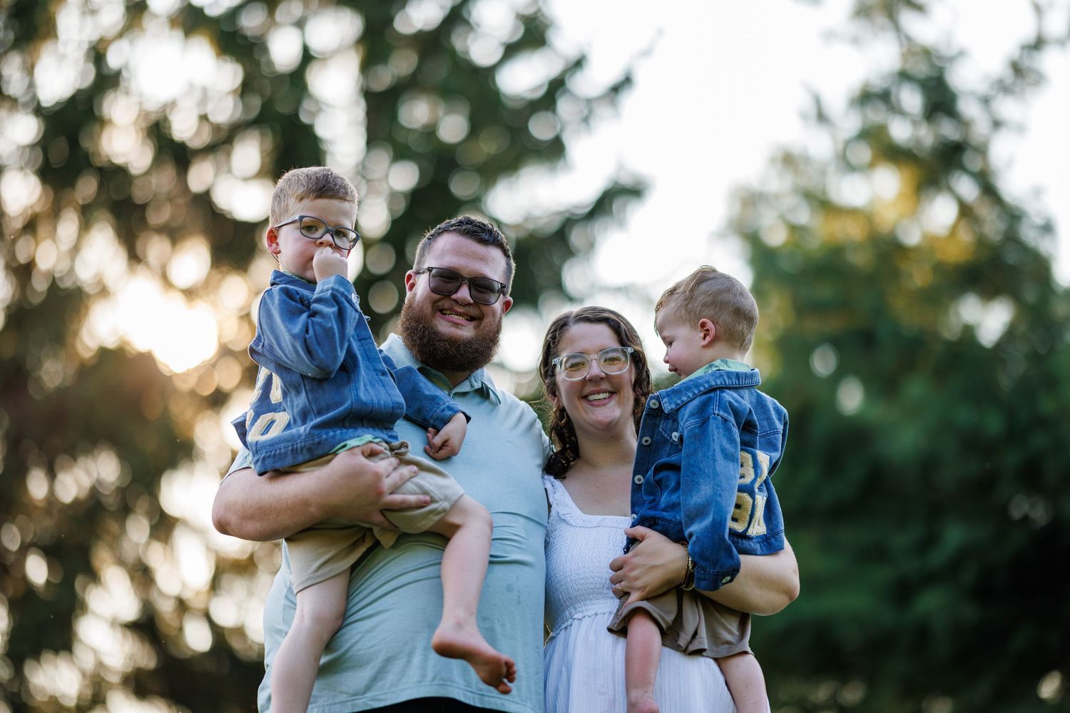 Happy couple holding two young boys outdoors with golden sunset light filtering through trees.