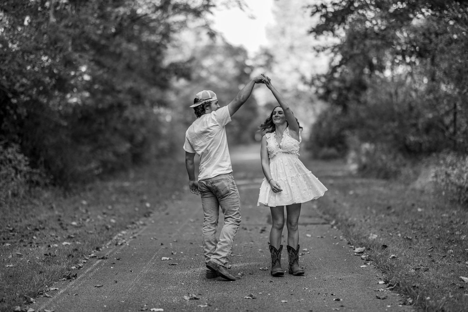 A black and white photo of a couple dancing on a tree-lined path, man spinning woman in a white dress.
