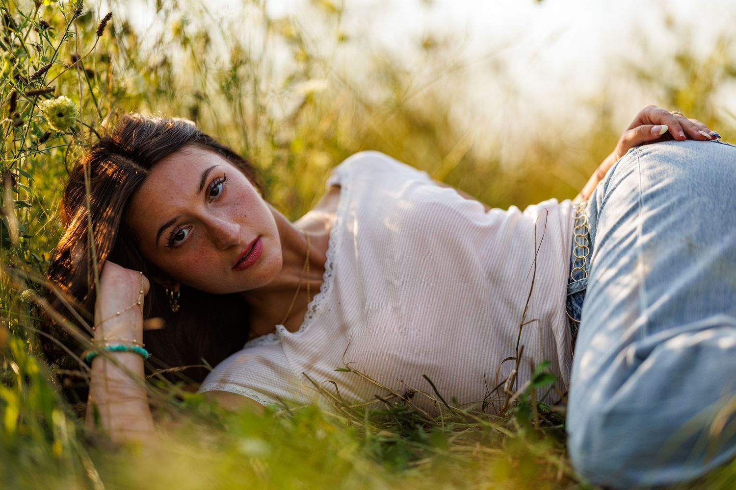 Young woman in white top and jeans lying in tall grass field during golden hour.