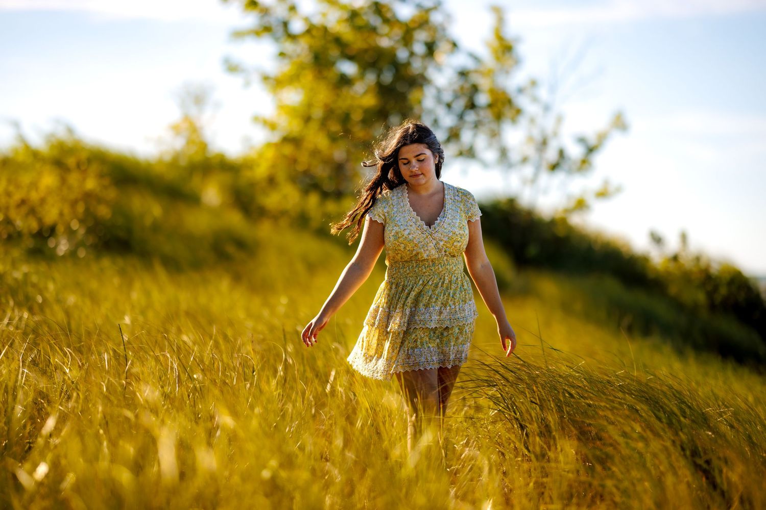 Woman in yellow floral dress walking through tall golden grass field at sunset.