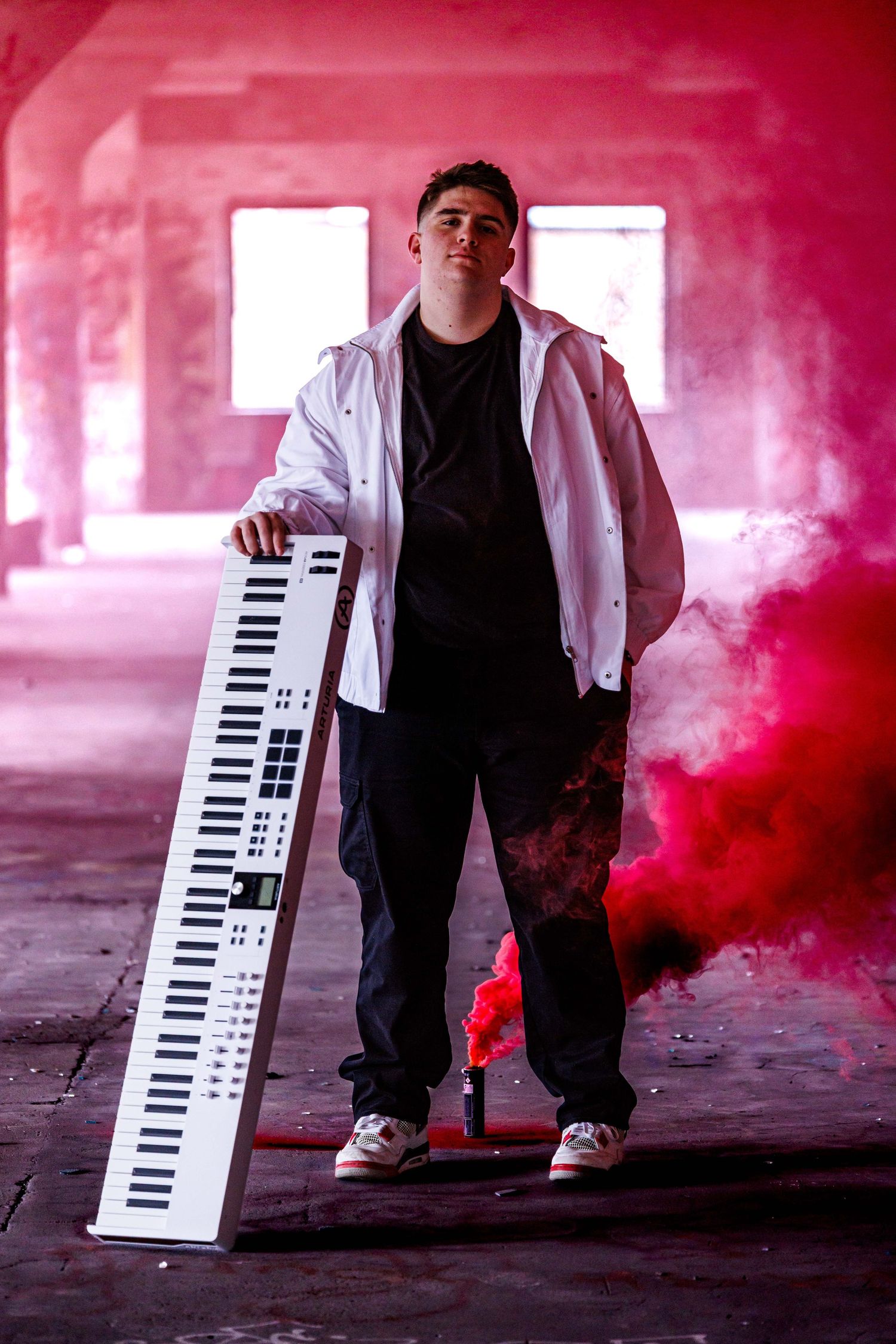 Musician in white jacket poses with keyboard amid red smoke in abandoned building.
