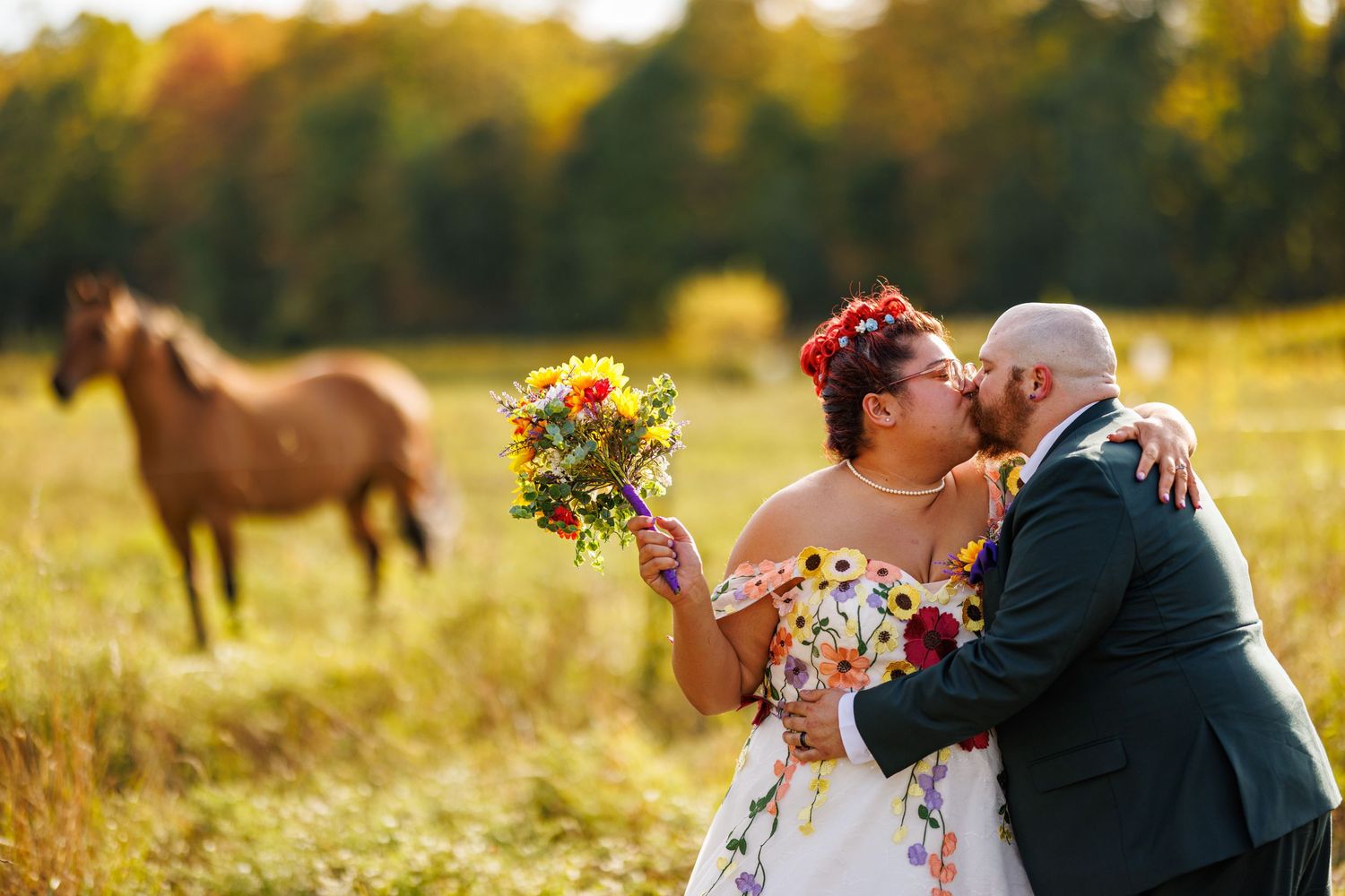 Bride and groom sharing a kiss in golden field with horse in background, bride holding wildflower bouquet.