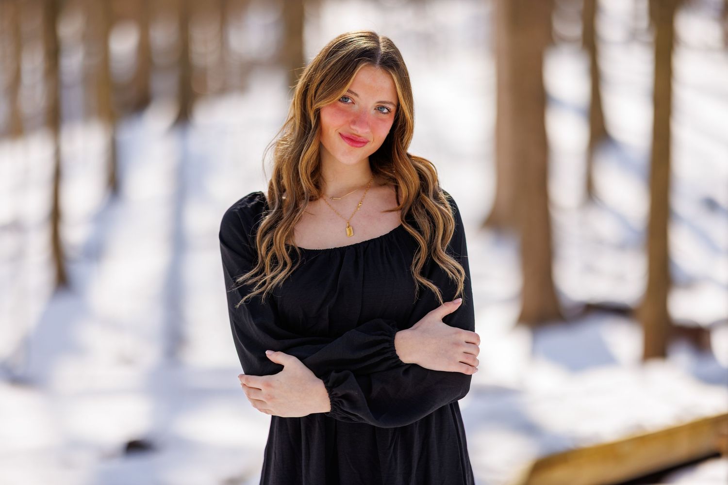 Young woman in black off-shoulder dress standing in a snowy forest with arms crossed.