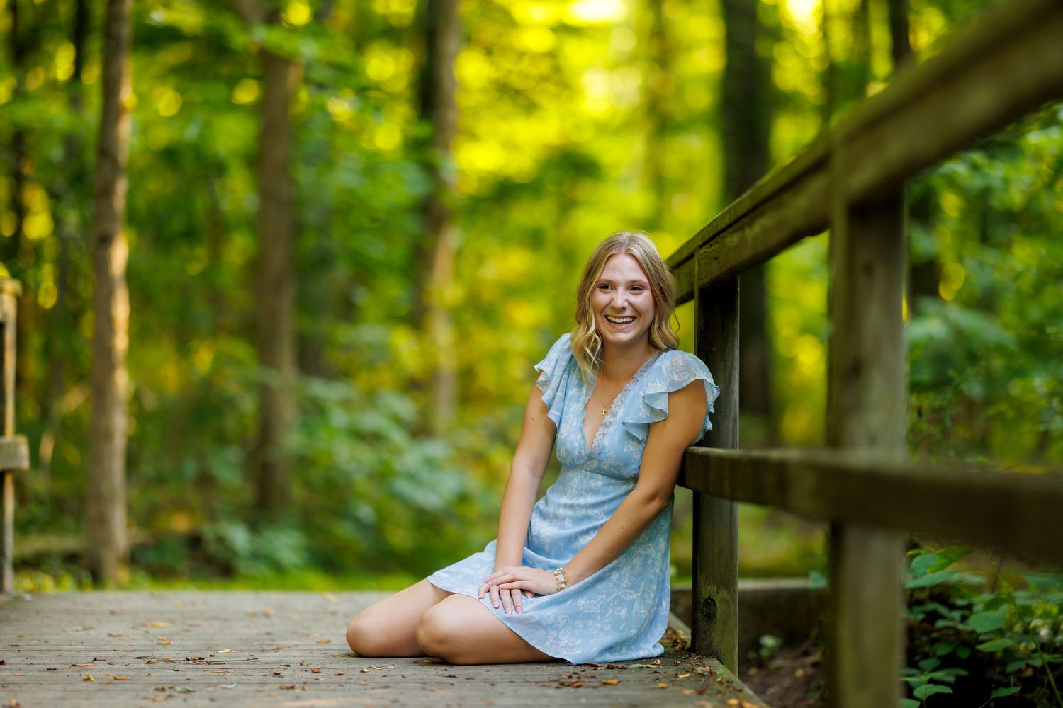 A smiling teenage girl in a blue dress sits on a wooden bridge in a forest.