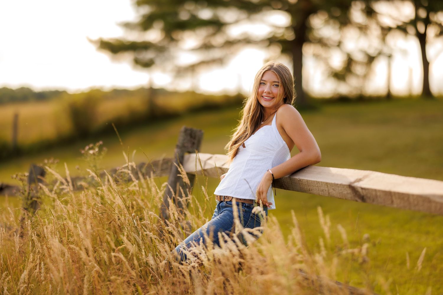 A teenage girl in a white top and jeans leans on a fence at golden hour.