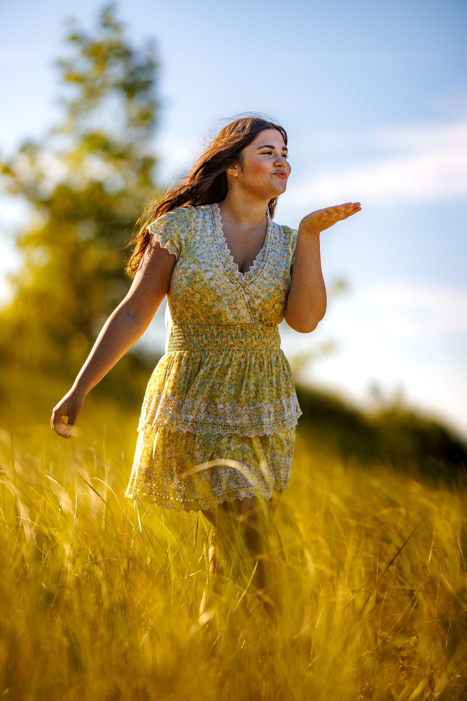 Happy young woman in yellow floral dress blowing a kiss while walking through a sunlit golden field.