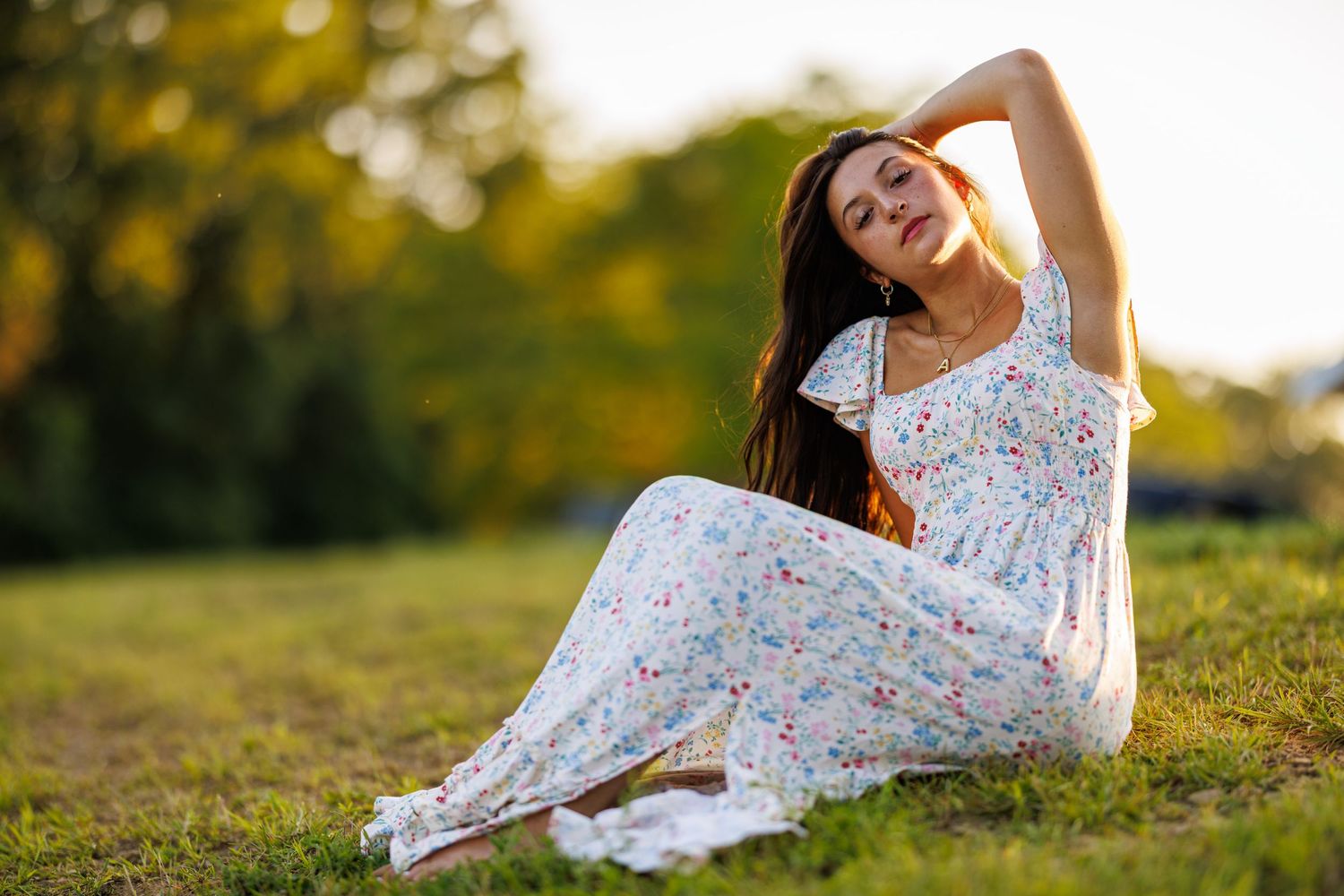 Woman in white floral maxi dress sitting on green grass during sunset.