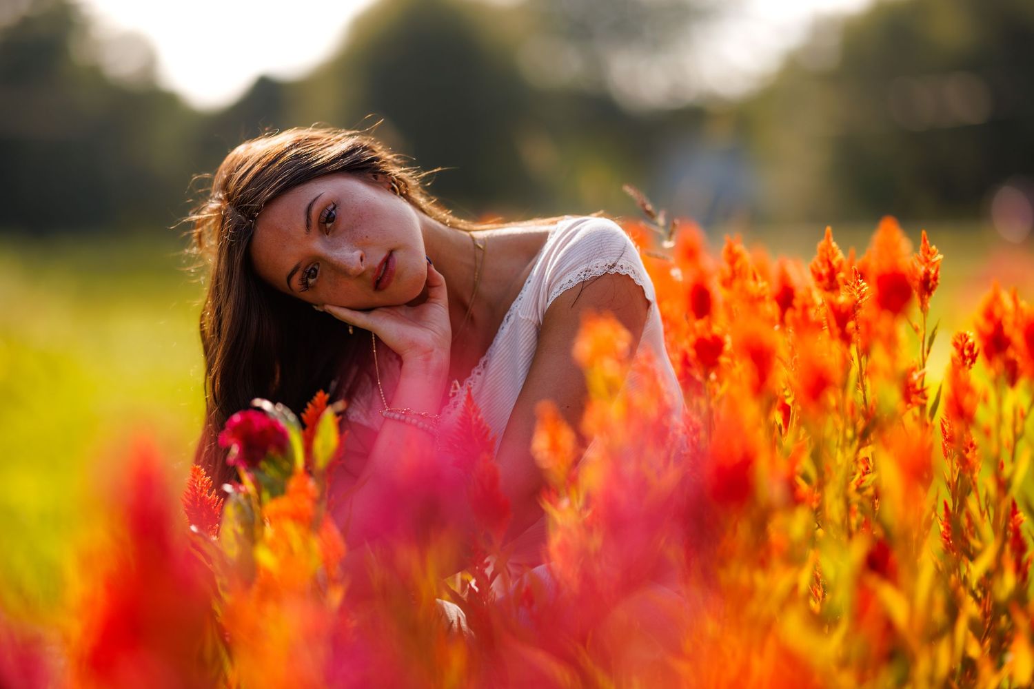 Young woman with long brown hair smiling among vibrant red and orange flowers in a sunlit field.