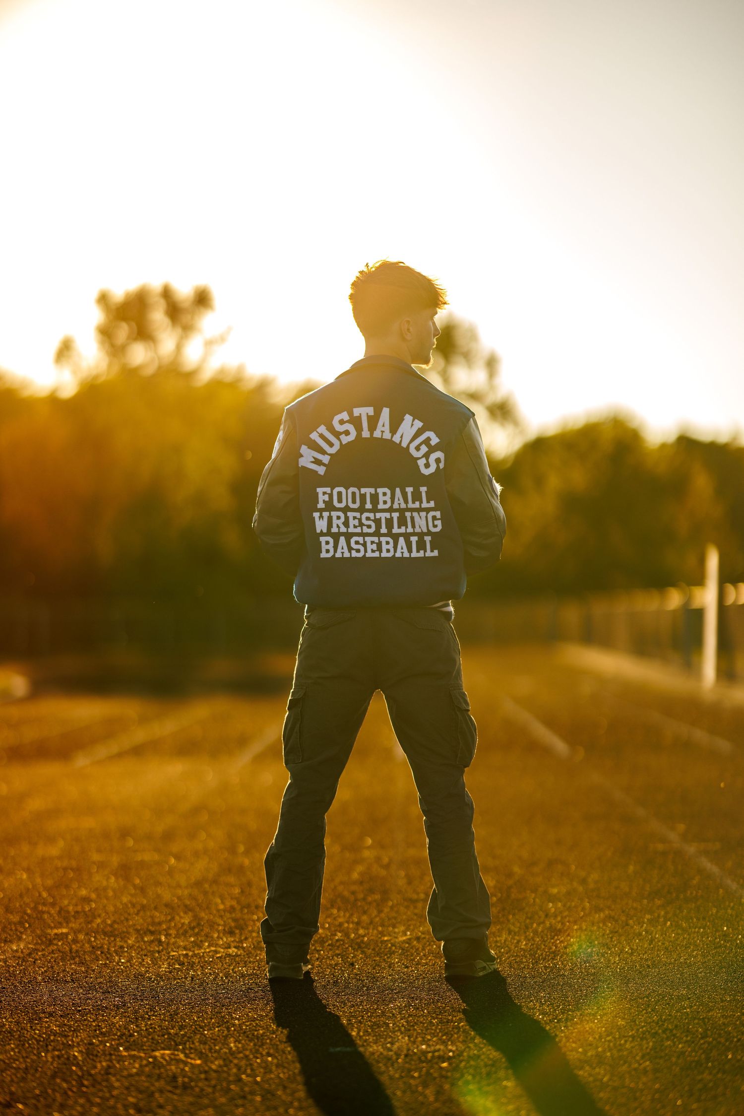 Teen boy in Mustangs football wrestling baseball jacket standing on track at golden sunset.