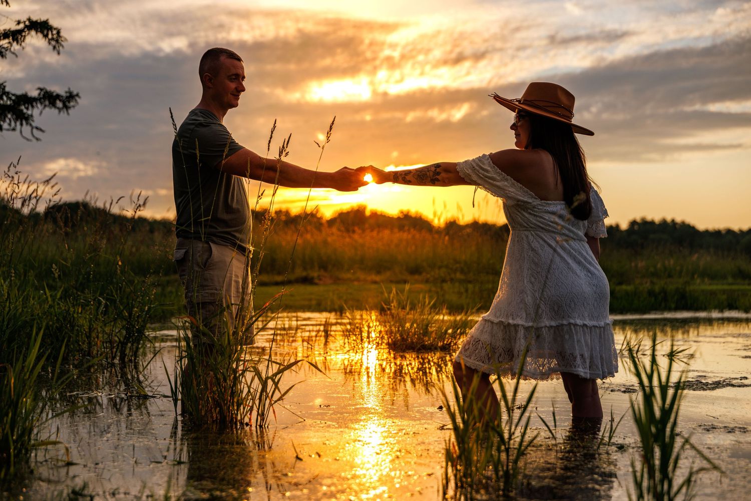 A couple holds hands while standing in water during a vibrant sunset in a marshland.