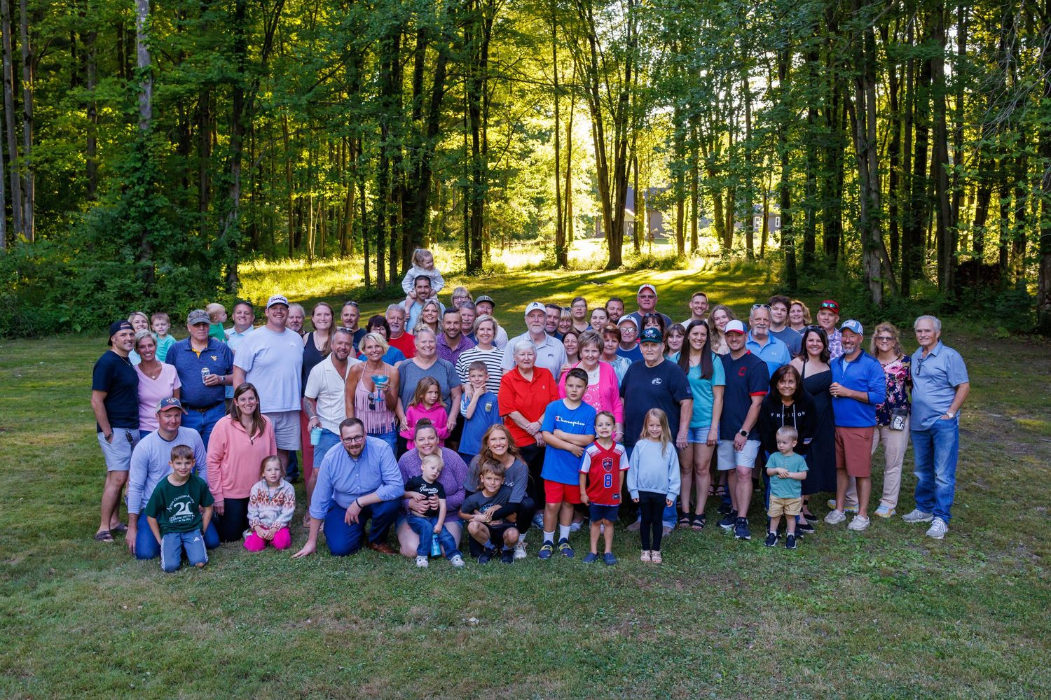 A large extended family gathers for a group photo on a lush green lawn in the woods.