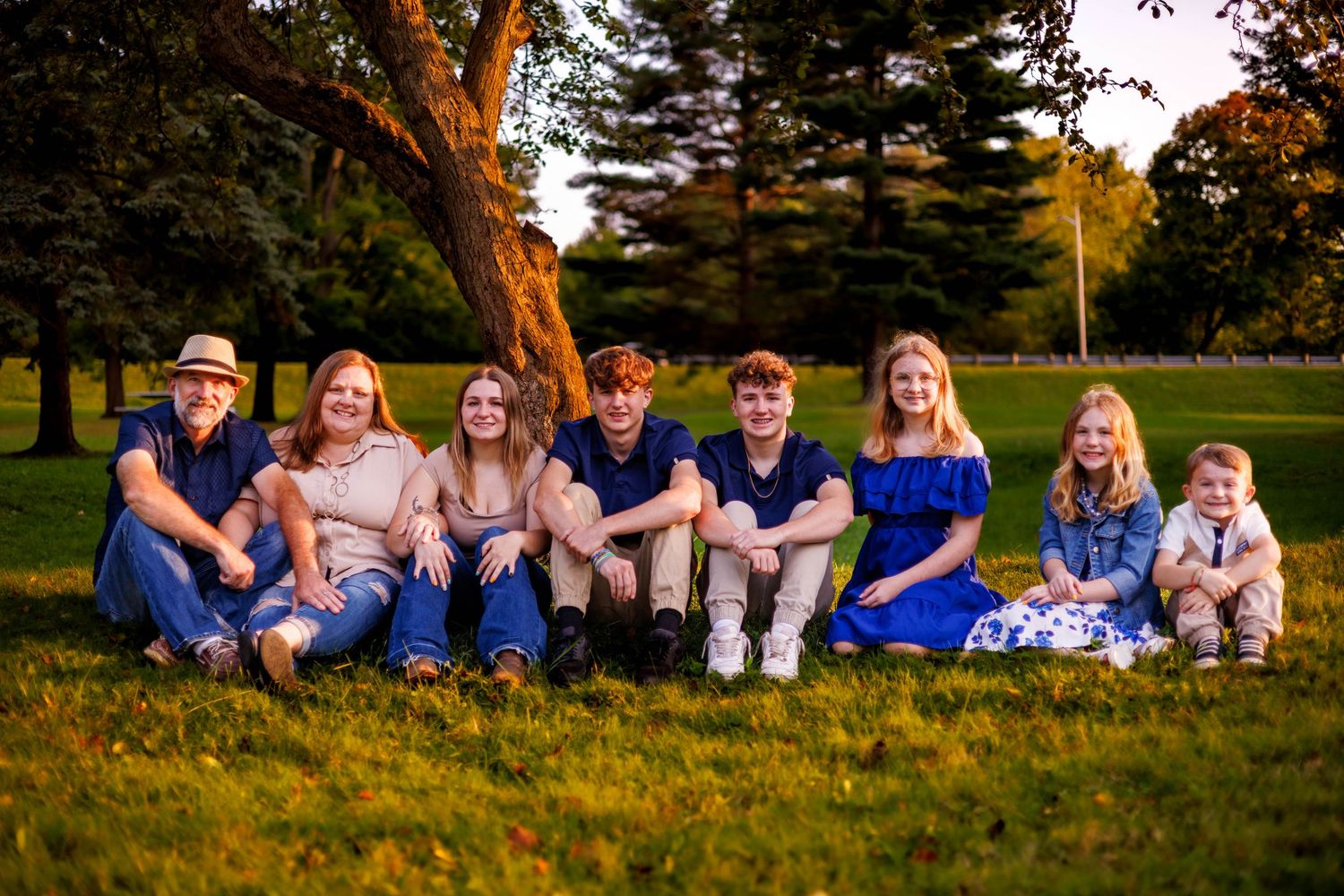 Extended family of eight sitting together on grass beneath a tree at golden hour.