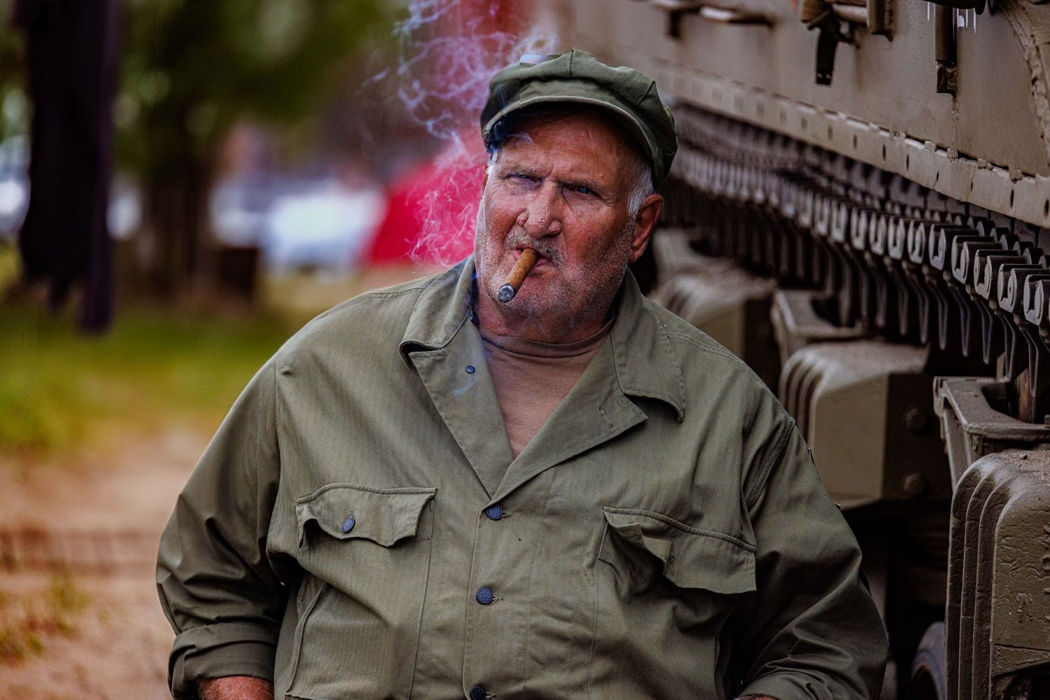Older man in olive military uniform smoking cigar leaning against armored vehicle outdoors.