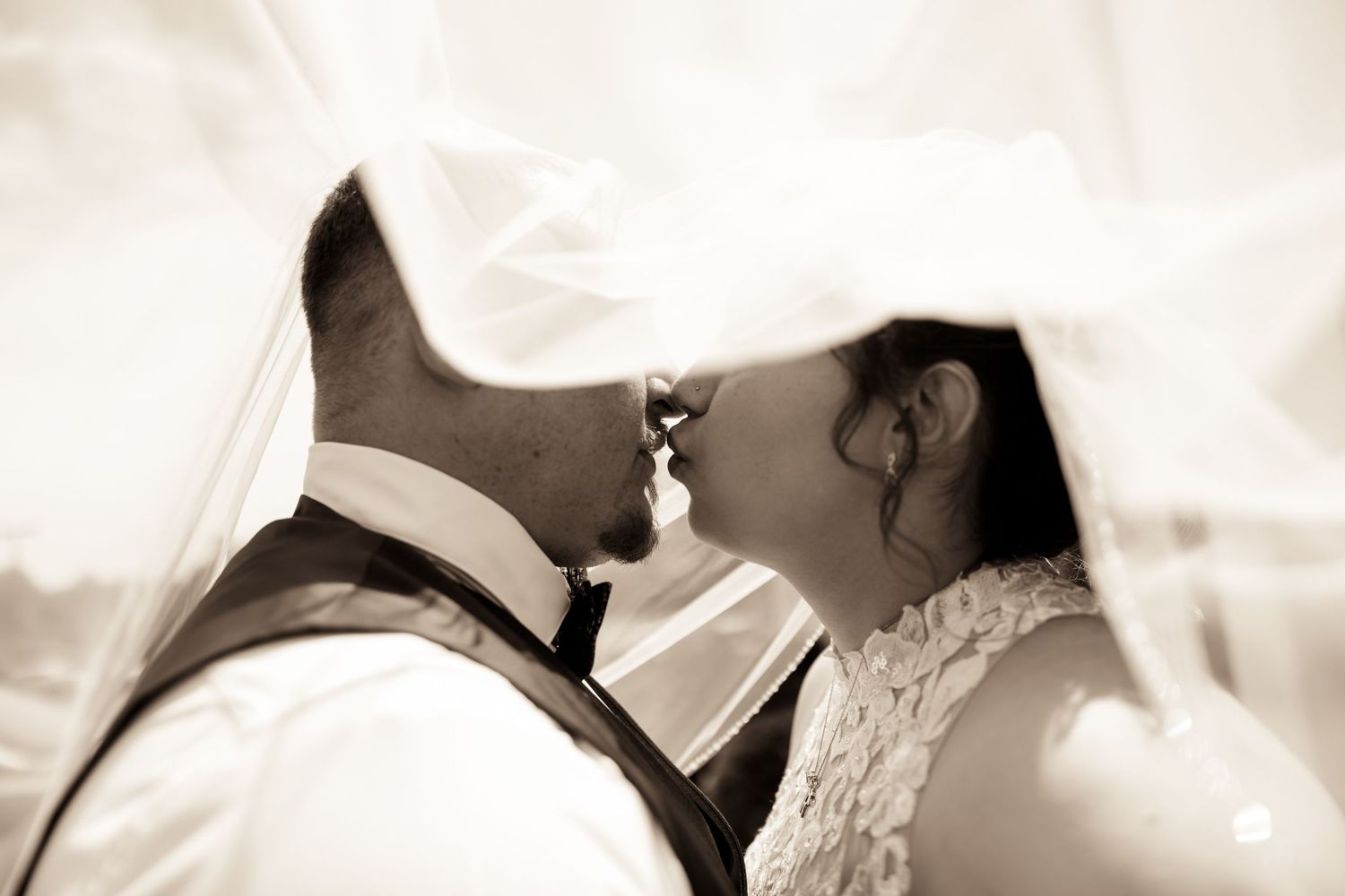 Bride and groom sharing a romantic kiss under a flowing veil in soft sepia-toned wedding photography.