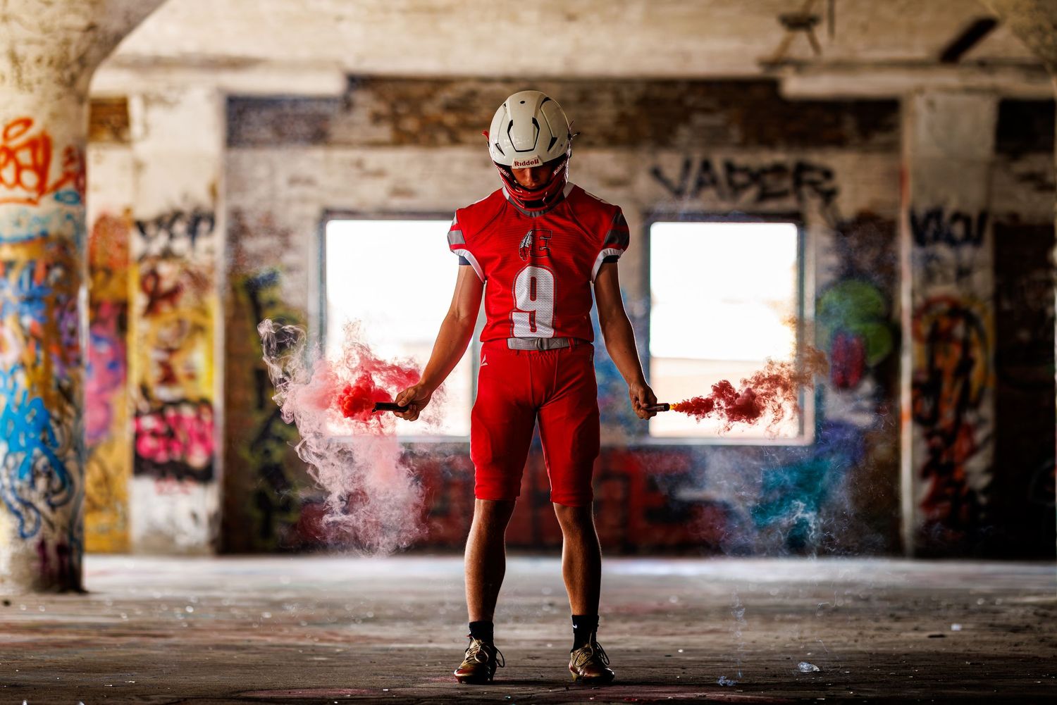 Football player in red uniform holding colorful smoke grenades in an abandoned graffiti-covered building.