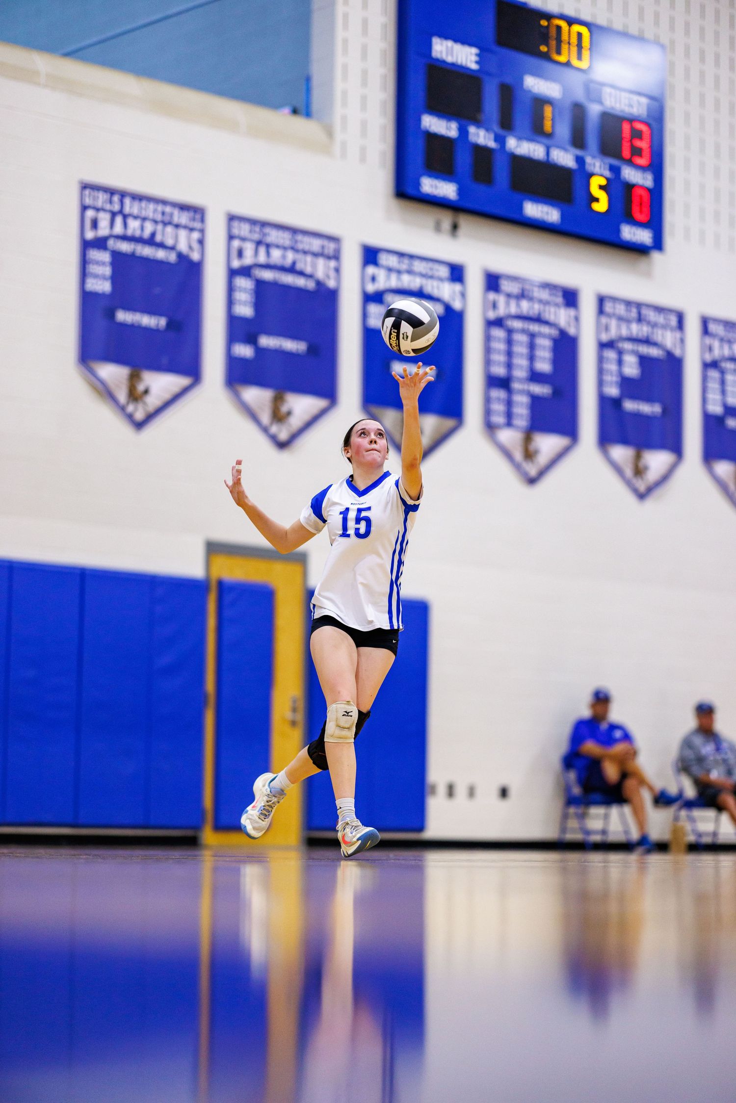 High school volleyball player number 15 serves the ball during an indoor gym game with scoreboard visible.