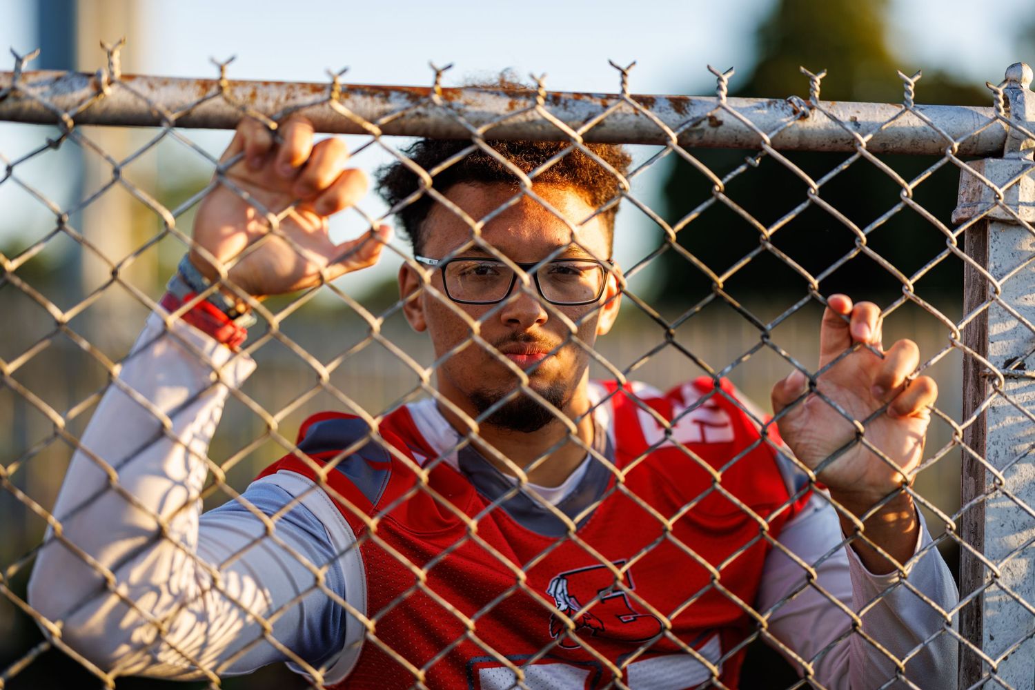 Young athlete in red jersey gripping chain-link fence outdoors during golden hour.