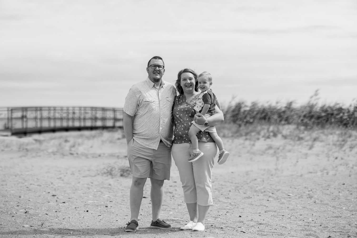 A smiling couple holds their young child on a sandy beach in a black and white photo.
