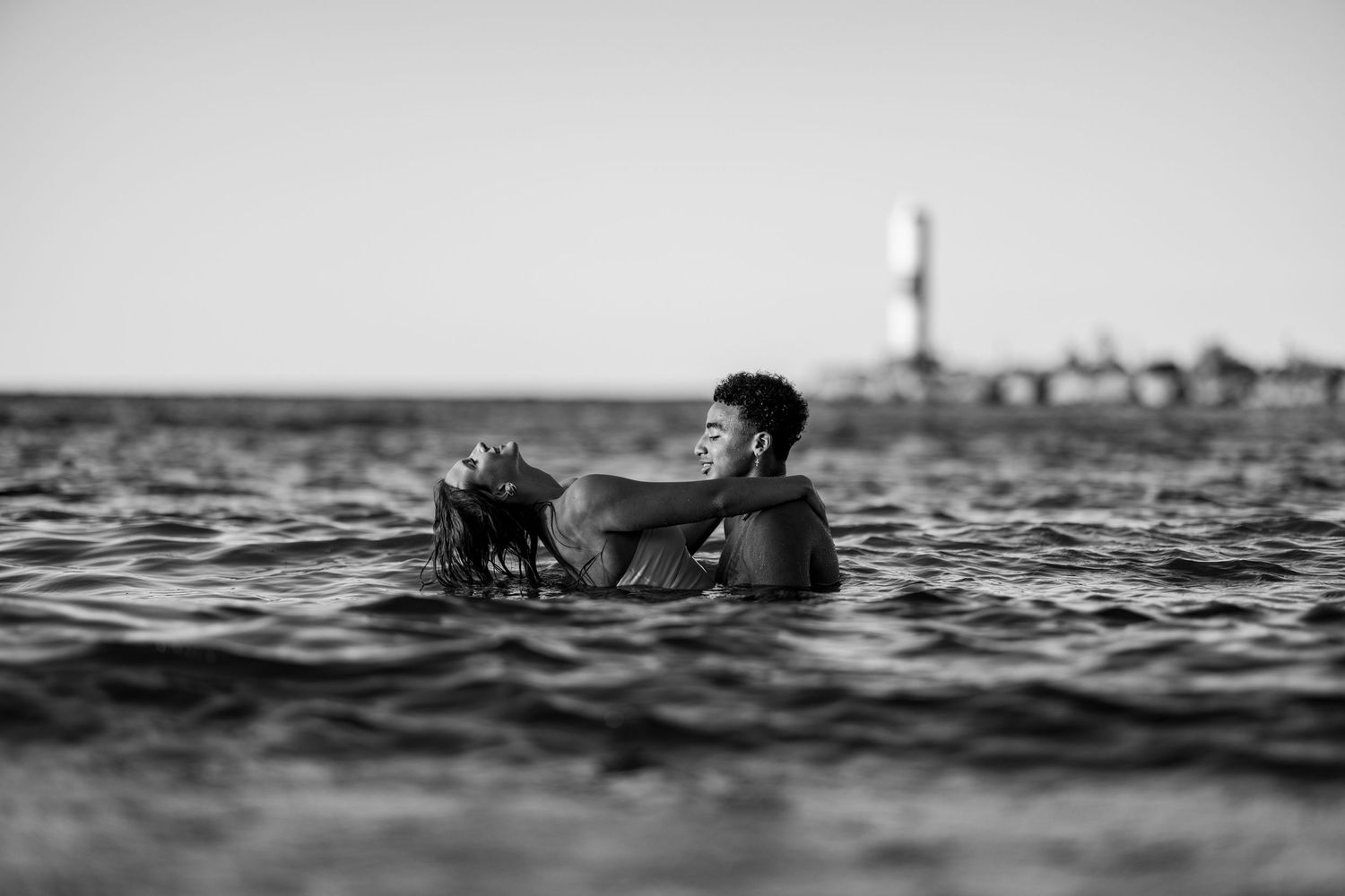A black and white photo of a couple embracing while swimming in the ocean with a lighthouse in distance.