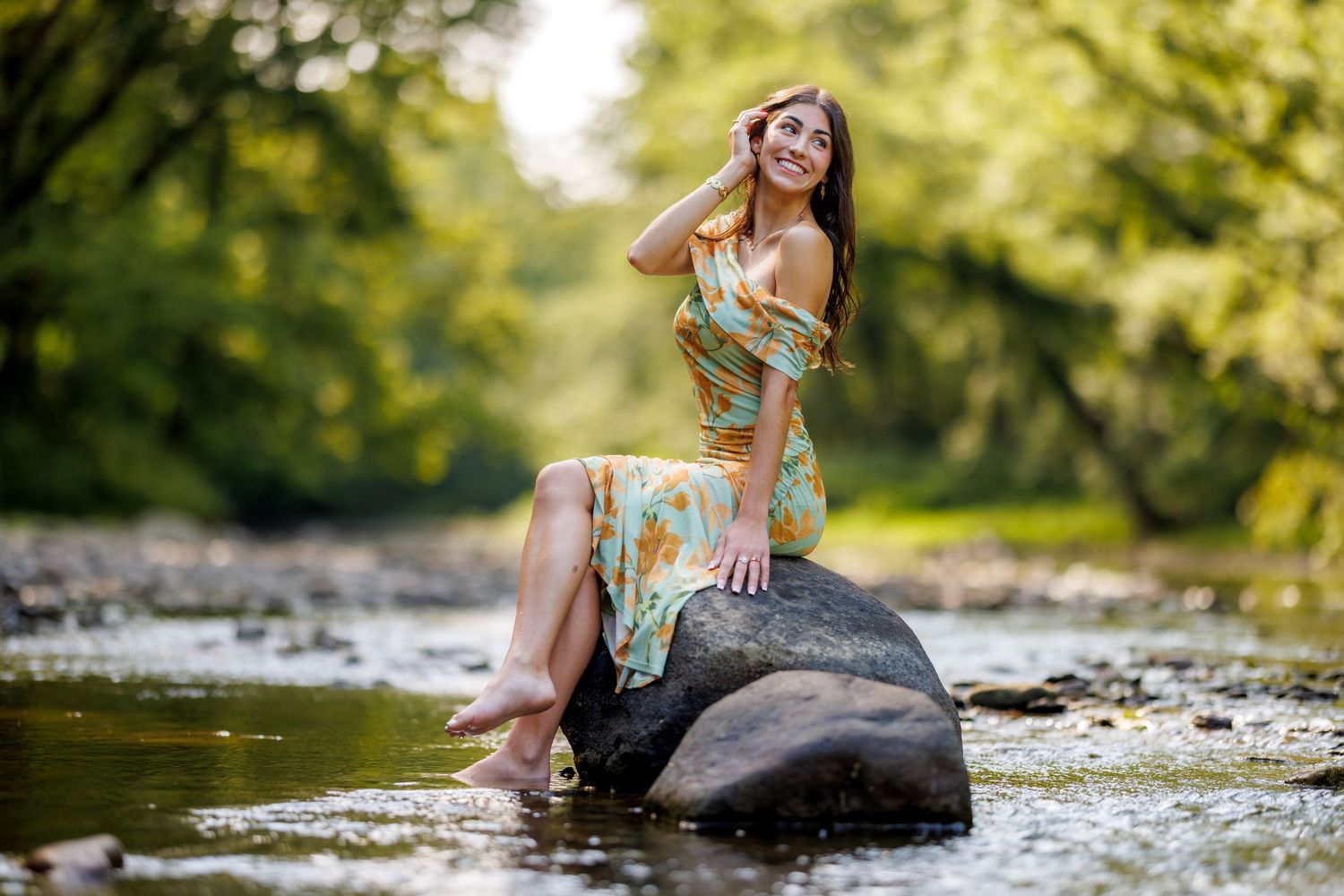 Smiling woman in a floral off-shoulder dress sitting on a rock in a shallow forest stream.