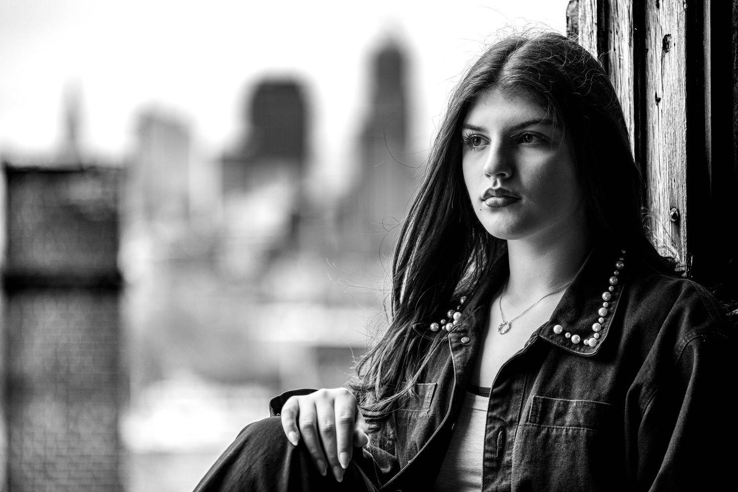 Black and white portrait of young woman with long dark hair sitting by window with blurred city skyline.