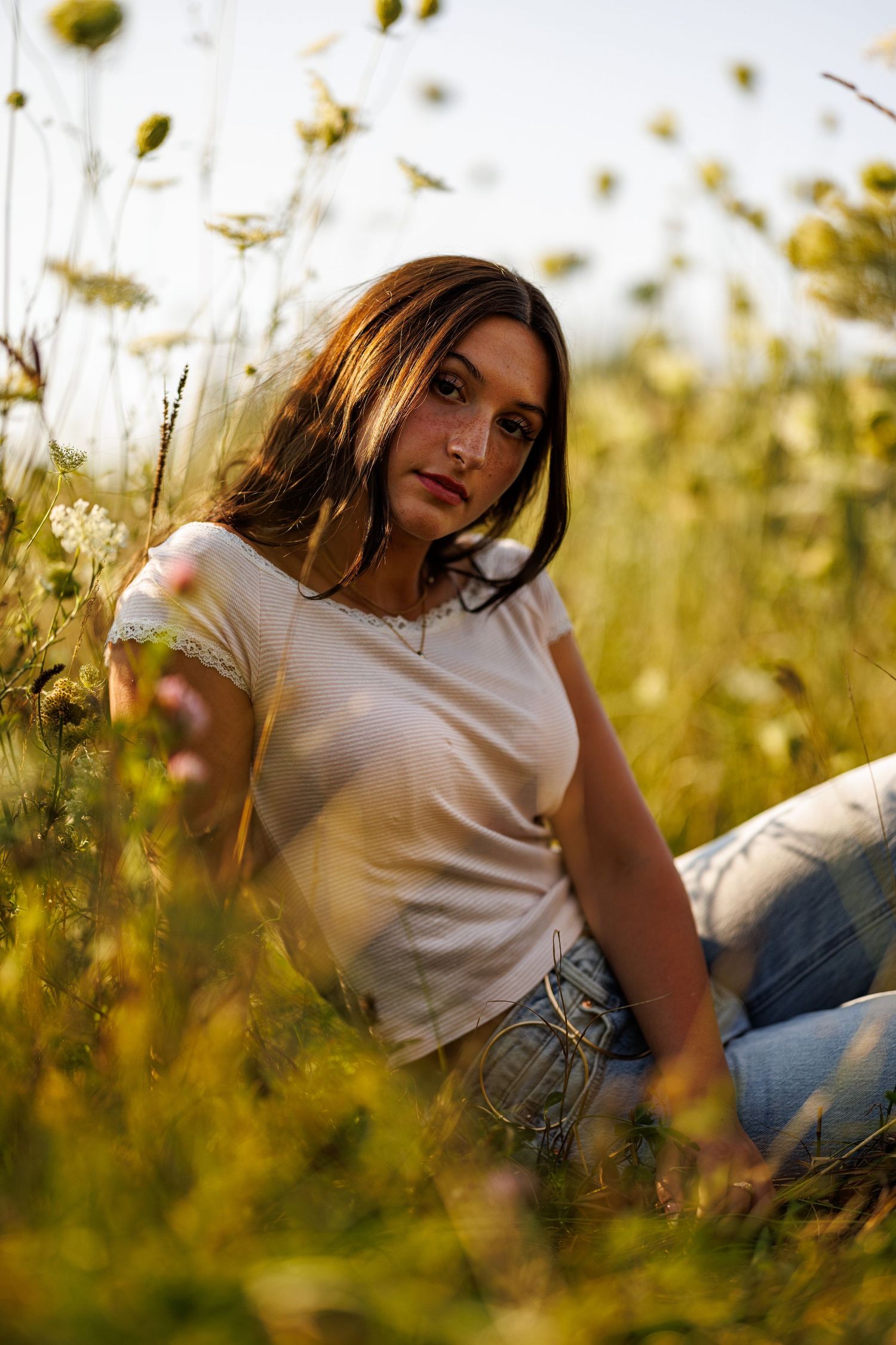 Young woman in white t-shirt sitting among tall green wildflowers in a sunlit meadow at golden hour.