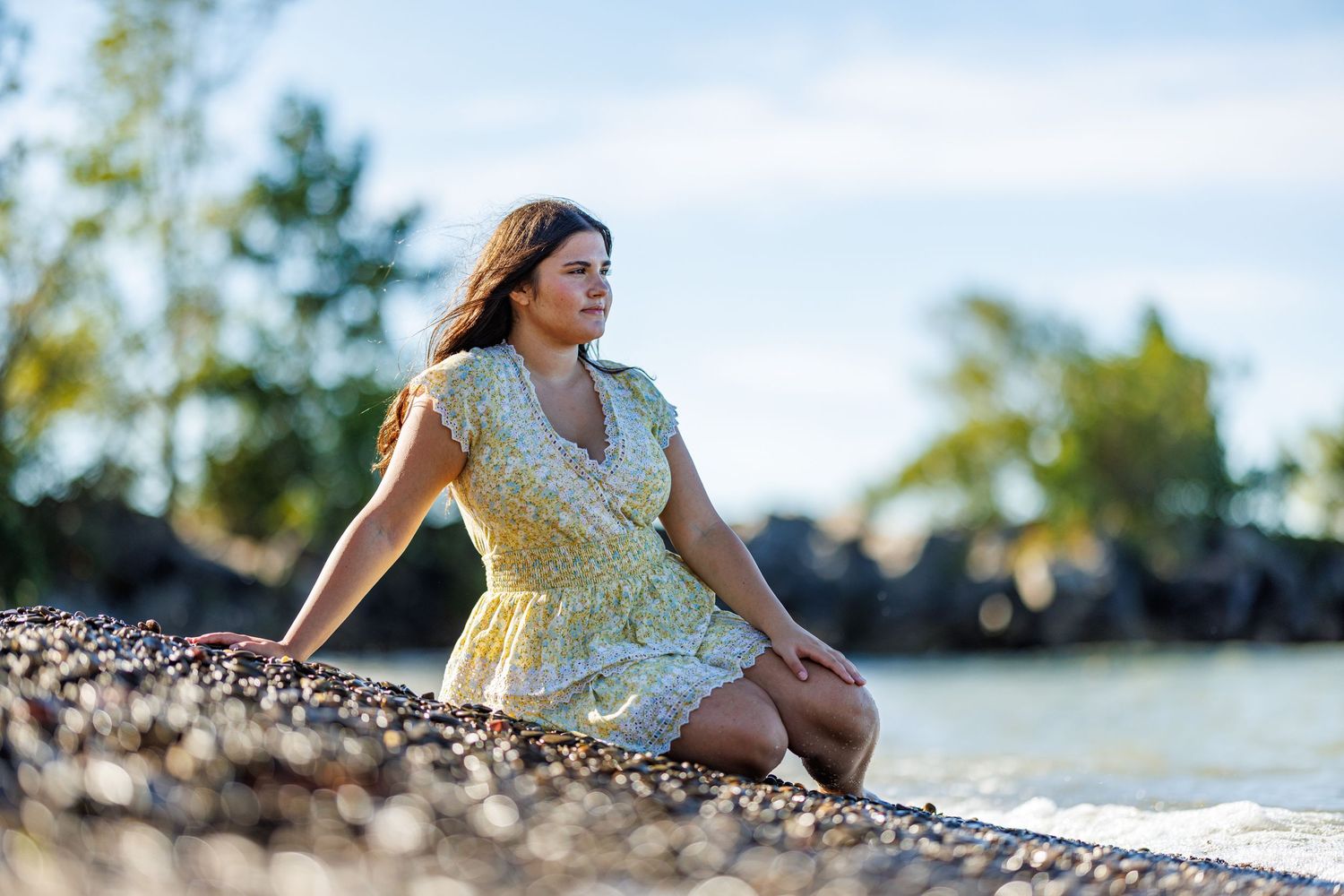Woman in yellow floral dress sitting on rocky shoreline gazing at the water.