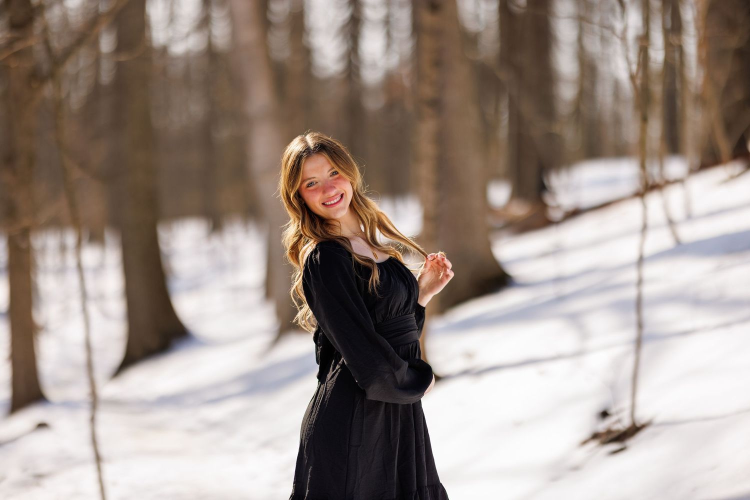 Smiling young woman in black dress standing in snowy winter forest with bare trees.