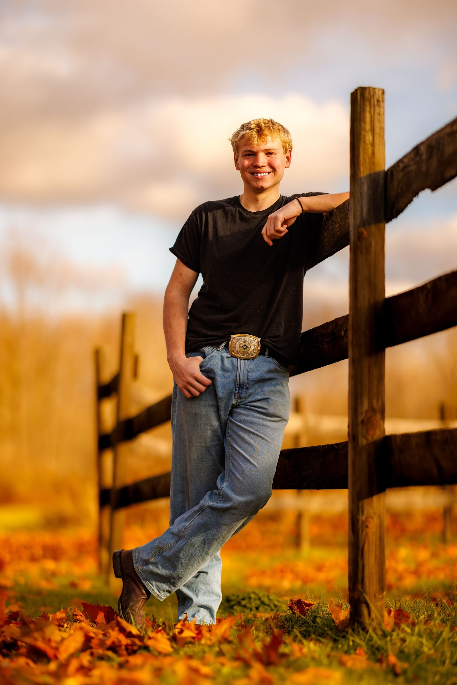 Smiling teenage boy leaning against a wooden fence in golden autumn light.