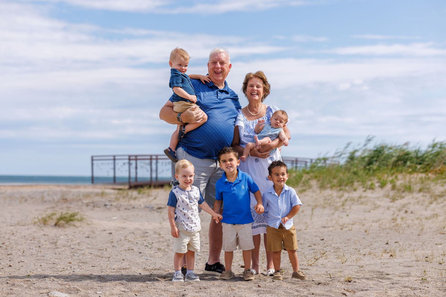 Grandparents pose proudly with four young grandchildren on a sunny sandy beach.