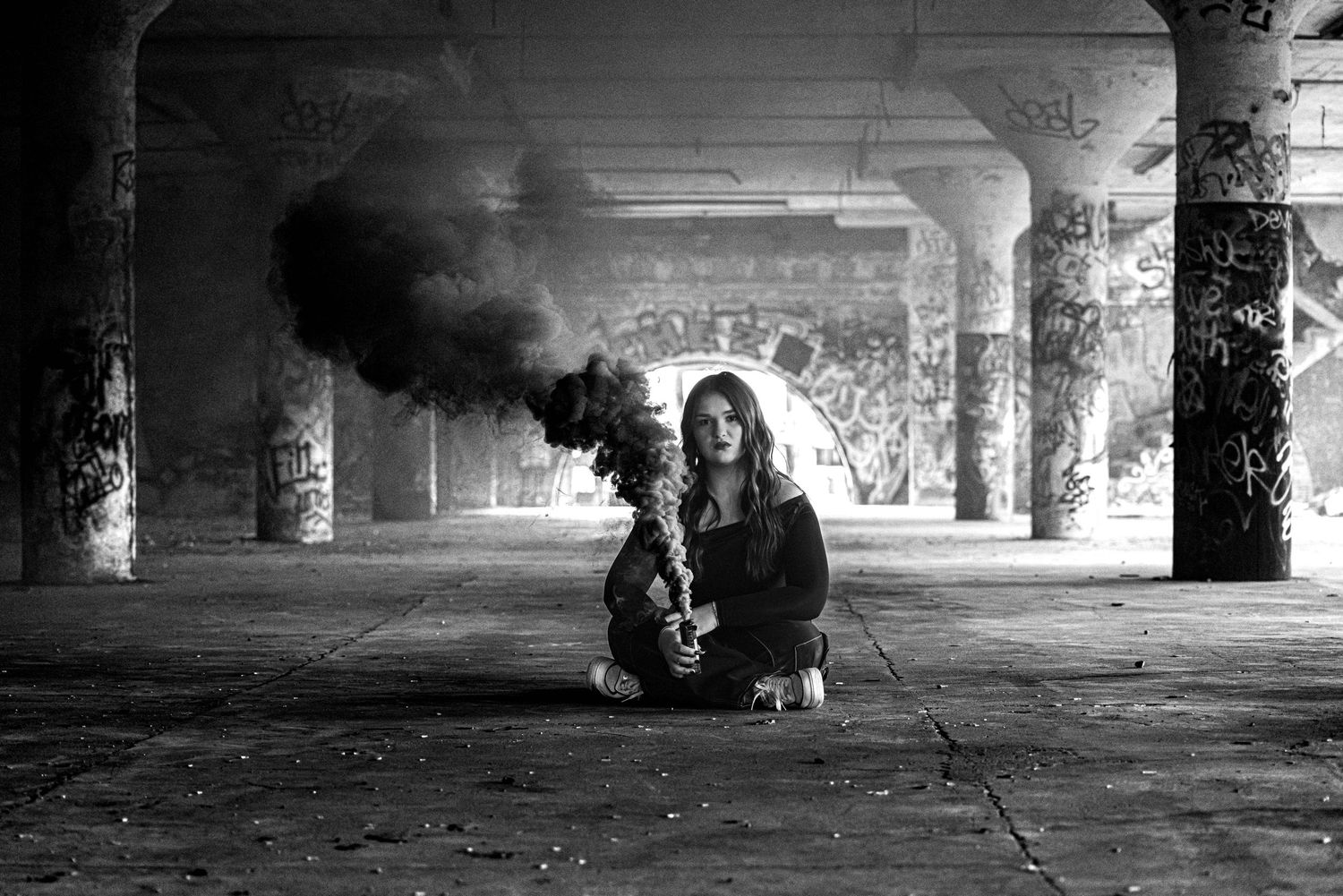 Woman sits with saxophone amid dark smoke in black and white abandoned warehouse photo.