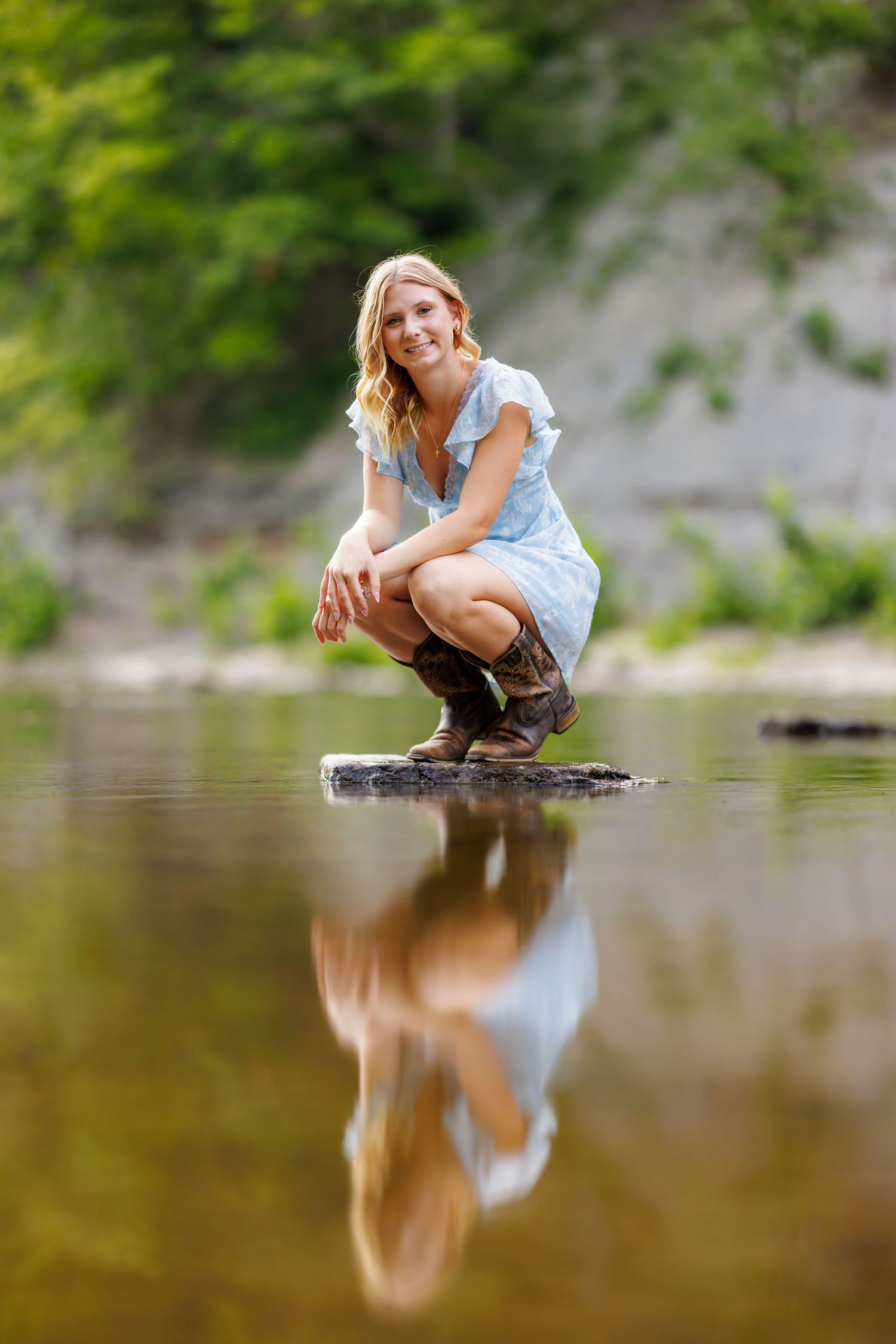 Smiling blonde woman in a light blue dress crouching on a rock in a calm river with reflection.
