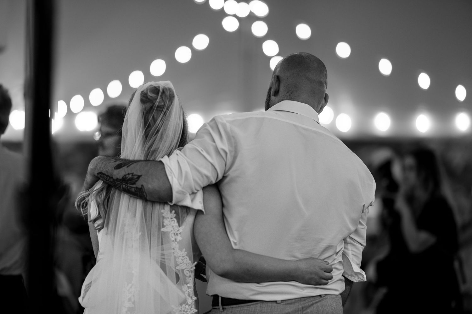 Black and white photo of a bride and groom sharing a romantic slow dance under string lights at their reception.