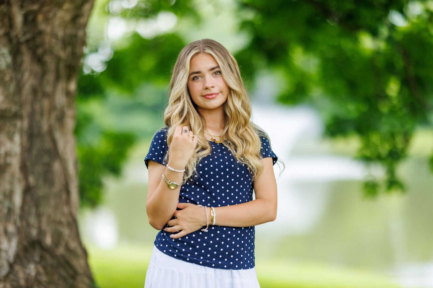 Blonde teenage girl in navy polka dot top posing near large tree for senior portrait outdoors.