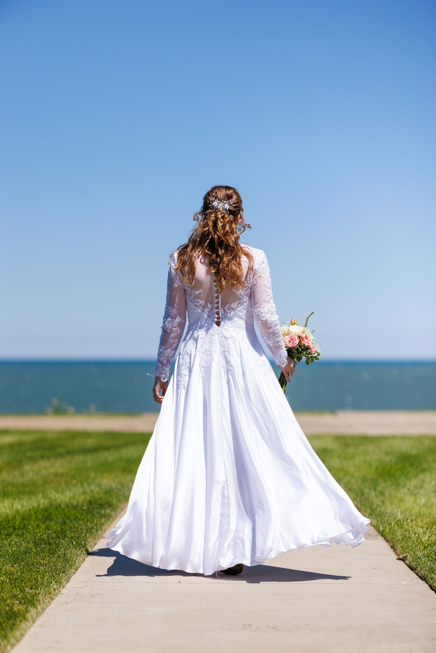 Bride in long-sleeved white lace wedding gown walking toward the ocean holding a colorful bouquet.
