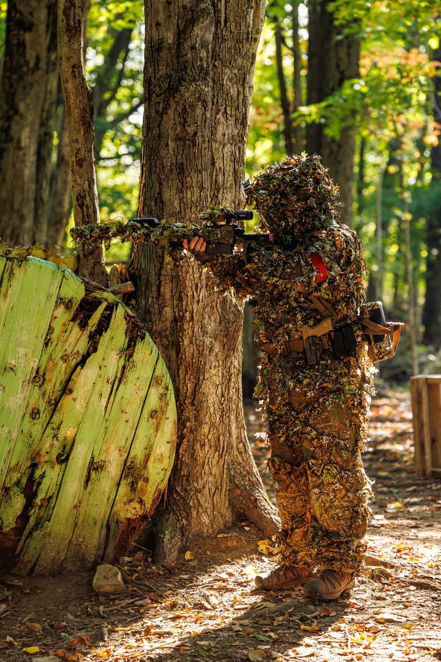 Camouflaged airsoft player aims rifle from behind a tree in a forest battlefield.