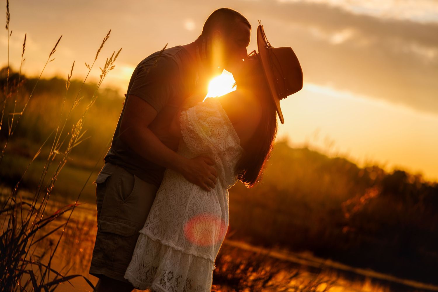 A couple shares a romantic kiss silhouetted against a warm golden sunset near a lake.