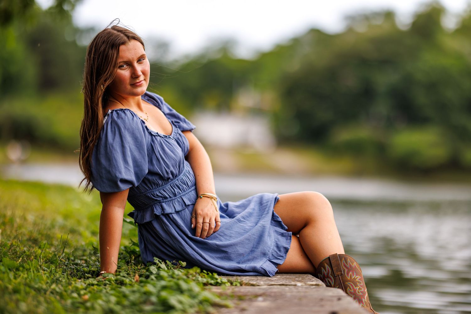 Smiling woman in blue ruffled dress sitting by lakeside wearing brown cowboy boots.