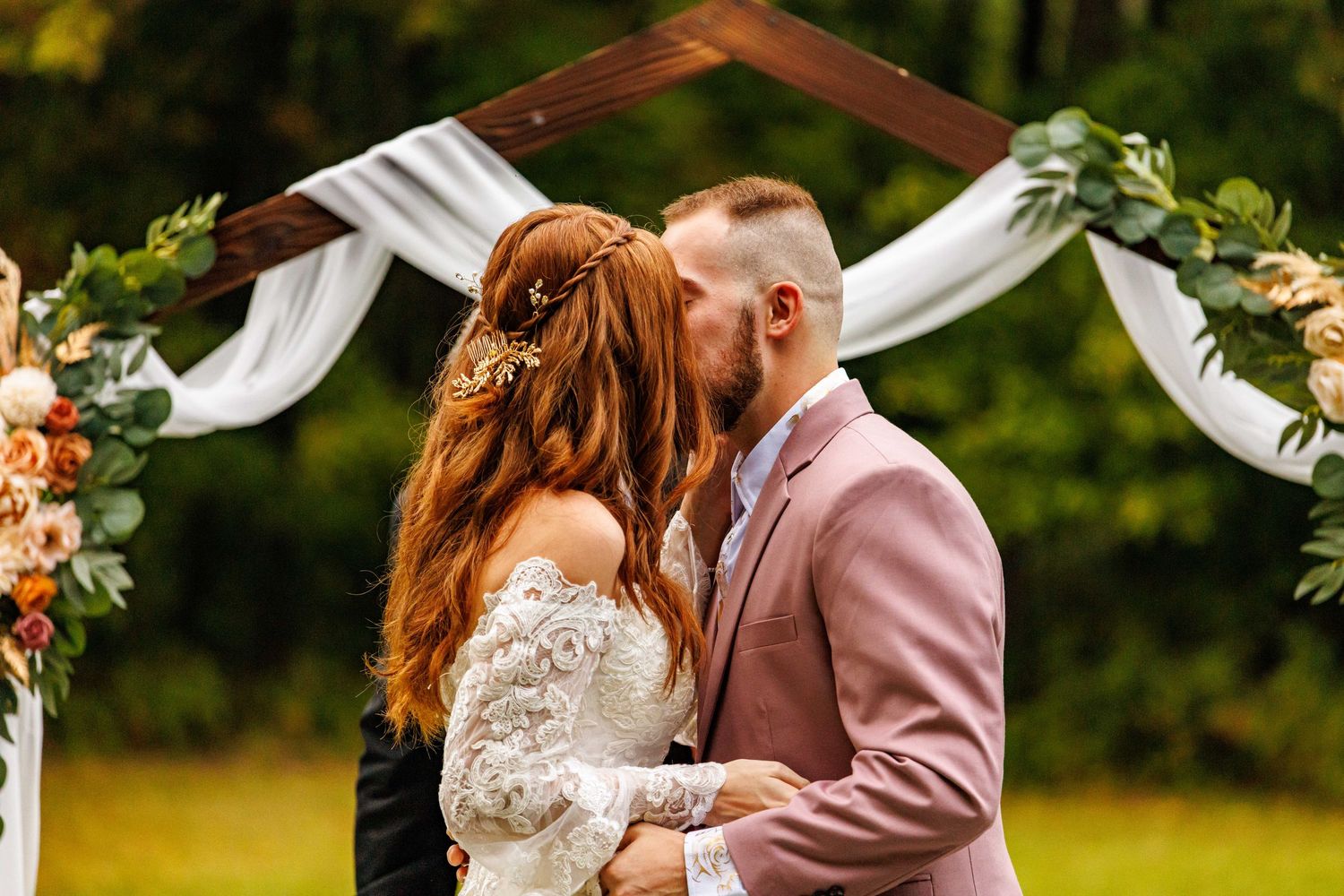 Couple kissing at outdoor wedding altar decorated with floral arrangements and white draped fabric.